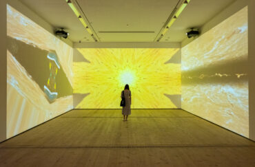 Patron Mono by Carolina Caycedo, image depicts a figure standing in a room with a three channel video emitting a yellow light