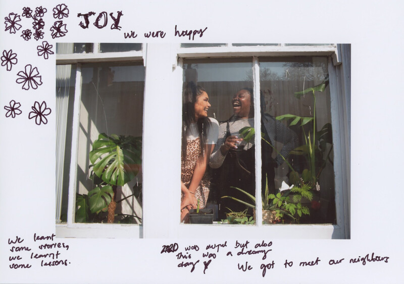 Two women laugh behind a window with plants in front of them 