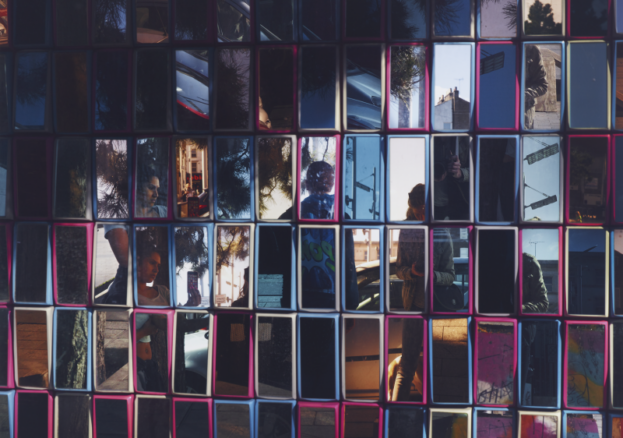 An artwork featuring many rectangular mirrors with coloured frames. In the mirrors are the fractured reflections of a young woman with dark hair, in the act of taking a photograph of two other unidentifiable young women in a street.