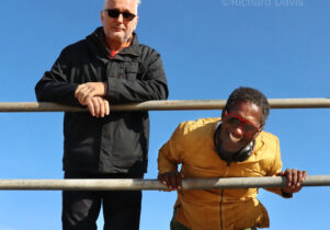 Two men stand at railings with blue sky behind. Both are wearing sunglasses and one is leaning forward with his head under the top railing and laughing. 
