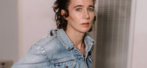 a white woman with swept back culy auburn hairs stares into the camera.  she is in a white room near an air conditioning vent and is wearing a faded denim jacket