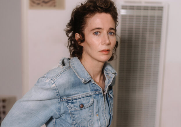 a white woman with swept back culy auburn hairs stares into the camera.  she is in a white room near an air conditioning vent and is wearing a faded denim jacket