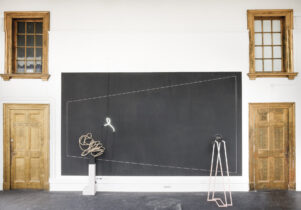 A minimalist art installation features geometric wire sculptures, a neon light in front of a black rectangle made of gesso, and two wooden doors with windows on either side in a white-walled room.