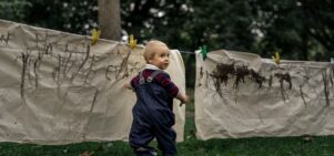 Toddler standing on grass with a paintbrush in hand turning to the right to look at the camera. He is wearing a navy blue rainsuit with a burgundy and navy blue long sleeved tiop underneath. He is standing in front of large cream coloured sheets that have been strung up on a washing line. The sheets have mud marks making on.