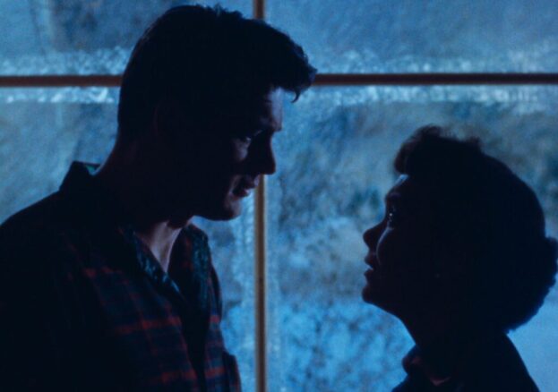 A man and a woman stood in front of a window at night look into each others' eyes  