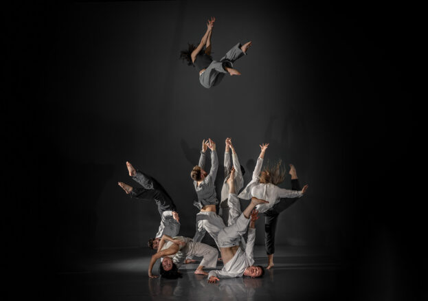 Hidden at the Lawrence Batley Theatre - A dynamic performance by a group of contemporary dancers from Motionhouse, showcasing acrobatic choreography. One dancer is mid-air in a dramatic flip, while others form intricate, intertwined shapes on the ground, reaching upward.