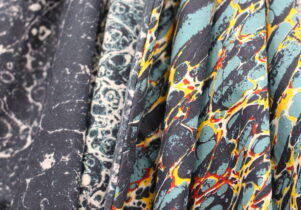 This image features a close-up of fabric with intricate, marbled patterns in various colors. The patterns resemble the swirling designs found in traditional marbling art. The fabrics display a range of tones, including dark greys, blues, teals, yellows, and red accents, creating a vibrant and textured appearance. The folds in the fabric highlight the material's flow and emphasize the rich detail of the designs.