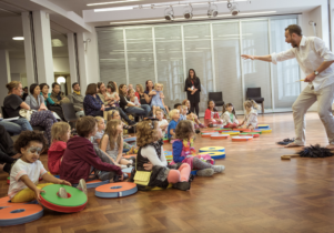A photo taken in an event space in Central Library, in which a man performs for a group of families with young children, who are dressed in bright colours.