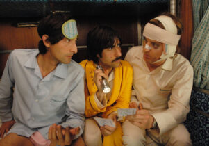 Three men sit next to each other. One's head is bandaged, one holds a torch and one wears a sleepmask.