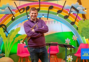 Sean Chandler stands in front of a rainbow with musical notes on it, holding a trumpet.
