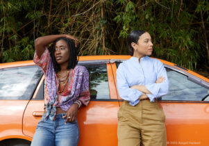 Two women stand next to an orange car.