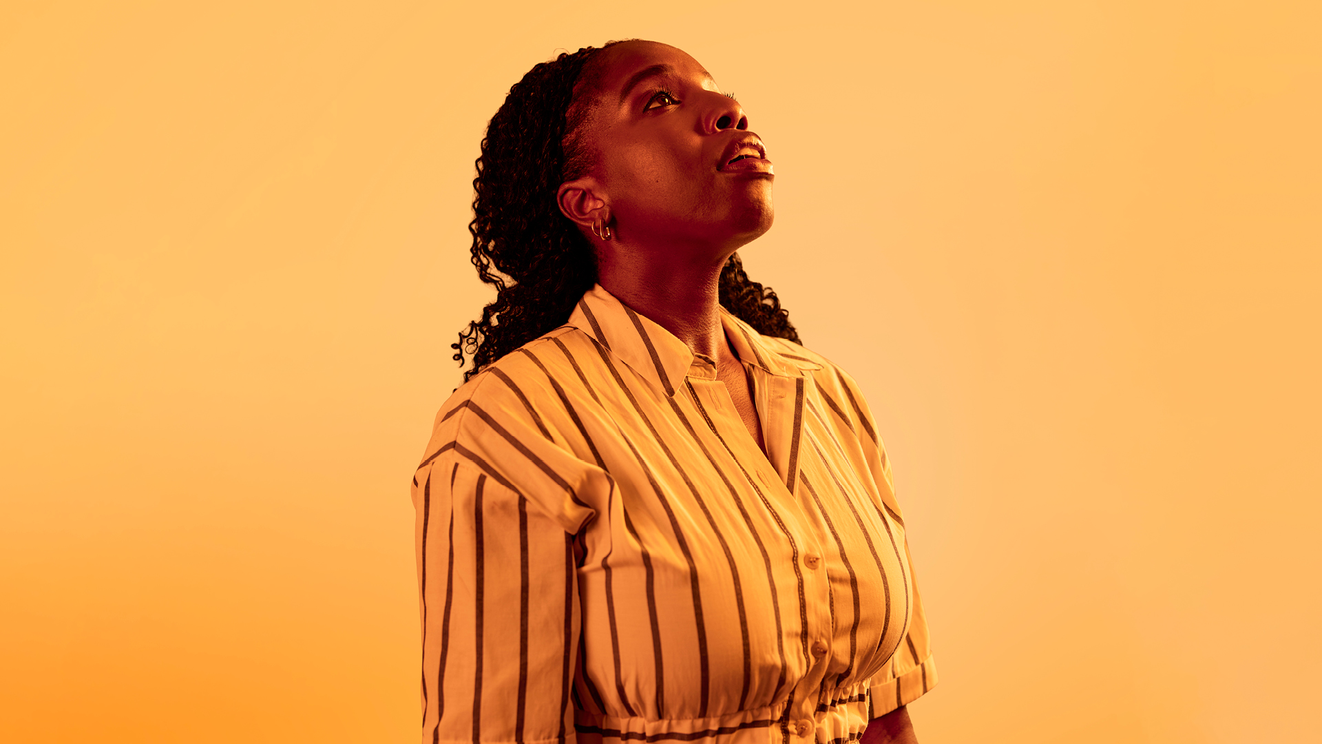 A woman of Black African heritage is drenched in yellow light. She looks up and away from the camera as if to a sunrise.