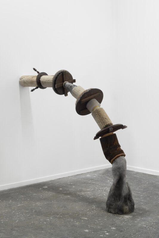 Abstract sculpture, tubular in form, bending from the floor to a wall somewhat like a pipe or a root. It is made in sections with different materials, including stone, iron, palm tree, wood salvaged from furniture, and miscellaneous pieces from abandoned factories.