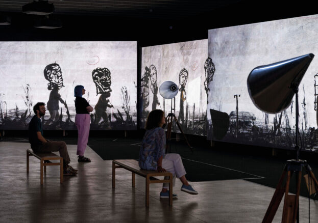 In a dimly lit gallery, three visitors observe large screens displaying monochrome animated figures. Two people sit on wooden benches, while a third stands, all captivated by the striking visuals. Studio lights illuminate the scene.