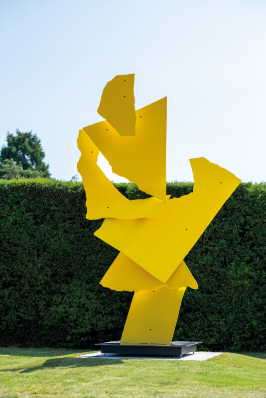 A large, abstract yellow sculpture stands prominently on a grassy area, surrounded by neatly trimmed hedges. The sculpture features dynamic, angular shapes that suggest movement and energy.