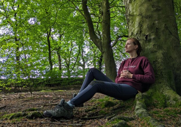 A lady sat at the base of a tree in a woodland holding a mug and looking reflectively