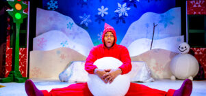 A performer in a bright red costume sits on a snowy stage set, holding a large snowball between their legs with a surprised expression. The colourful winter backdrop features snowflakes, hills, a snowman, and a traffic light with glowing lights.