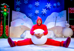 A performer in a bright red costume sits on a snowy stage set, holding a large snowball between their legs with a surprised expression. The colourful winter backdrop features snowflakes, hills, a snowman, and a traffic light with glowing lights.