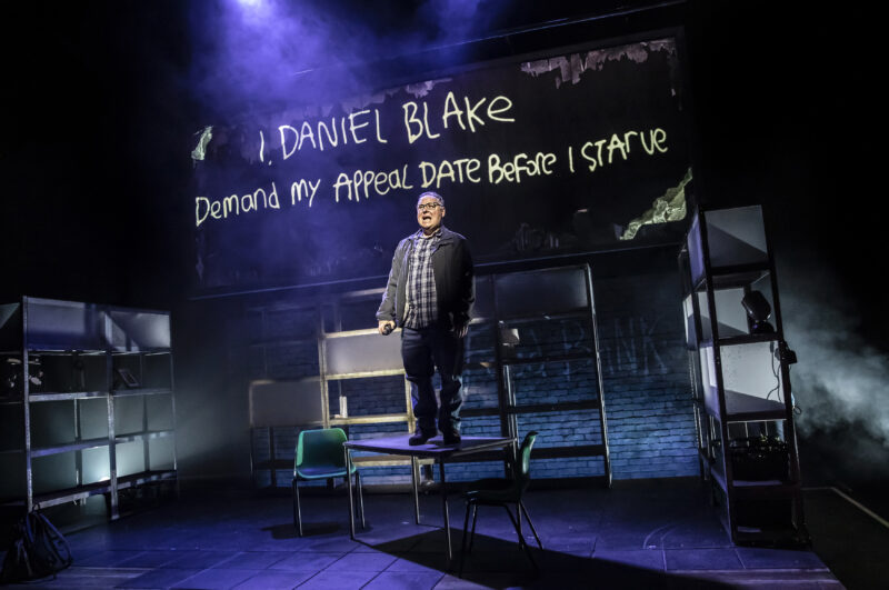An actor is performing on stage. They are standing on a table shouting with the words 'I, DANIEL BLAKE Demand my appeal date before I starve'.