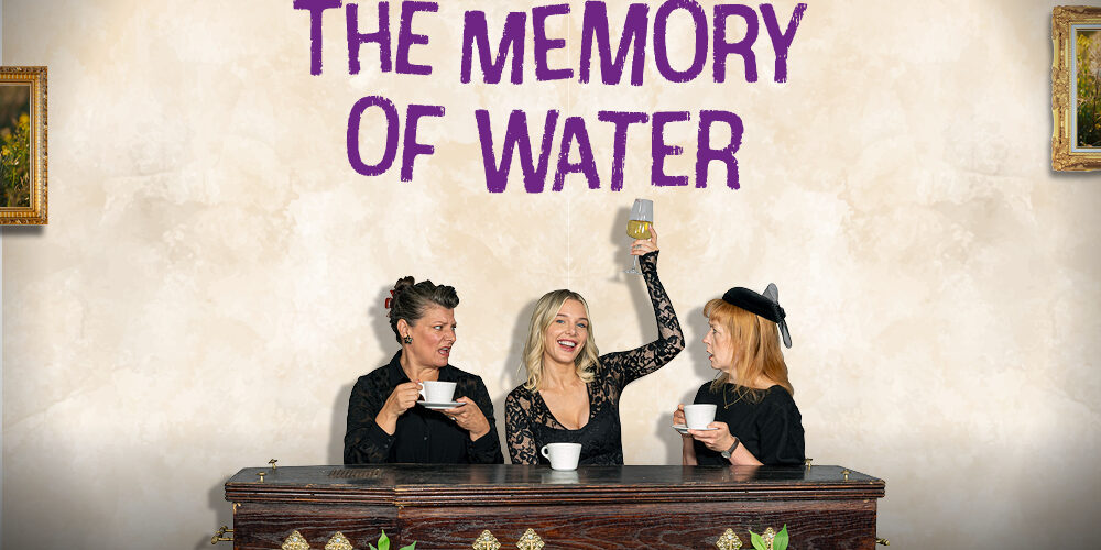 Text reads The Memory of Water. A image of three women in black sitting by a coffin, two with cups of tea, one in the middle raising a wineglass 