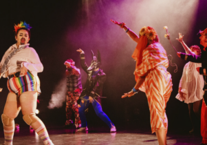 A number of performers dance on a stage wearing various costumes including rainbow hotpants and sunflower flower crowns