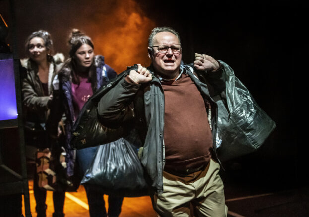 Three actors performing on stage, carrying black bin bags. 