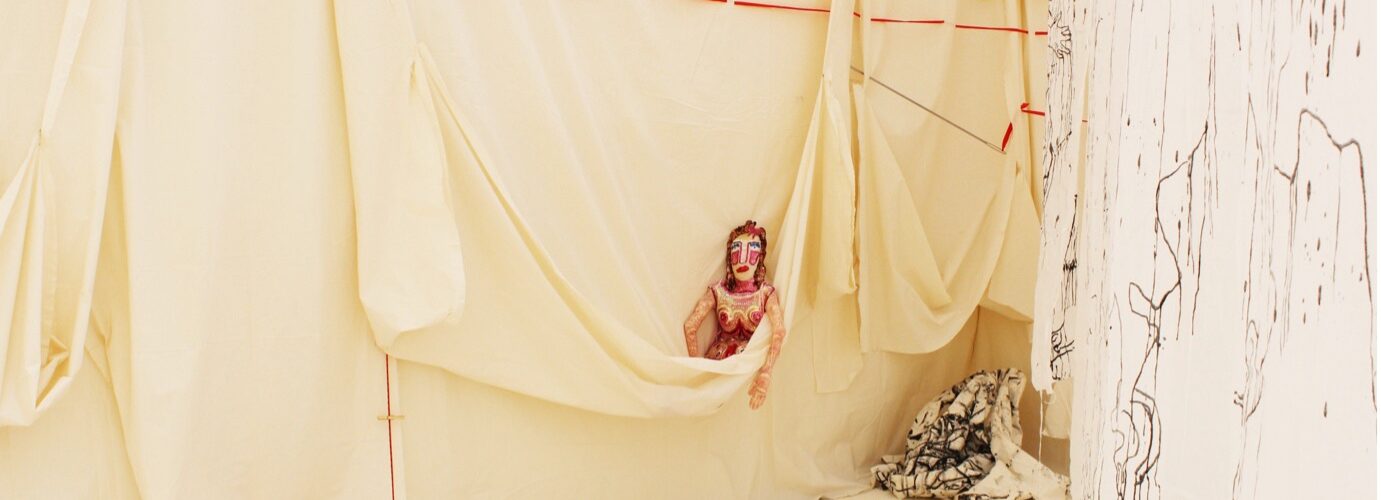 A doll with makeup peeks out of a hanging wall of butter yellow fabric. Red and black threads descend and cascade around the doll.