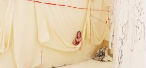 A doll with makeup peeks out of a hanging wall of butter yellow fabric. Red and black threads descend and cascade around the doll.