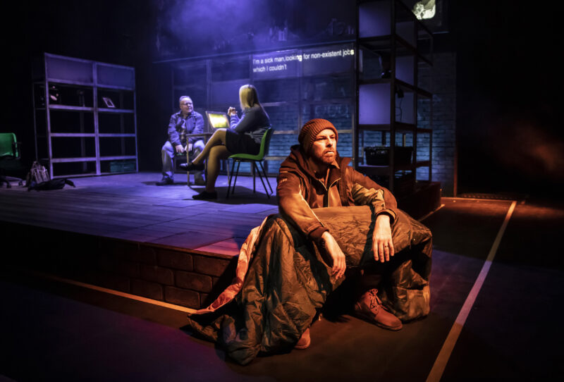 Three actors performing on stage. One is sat on the corner of the stage looking out.