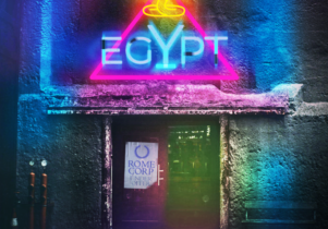 An image of a night club door with a pink and blue neon sign above it. The sign says Egypt with a gold neon cobra above it. On the club door is a sign saying Rome Corp - Under Offer