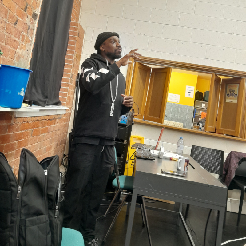 A man can be seen standing by a table, he holds his right arm up as if he is giving some direction to the actors in the rehearsal.