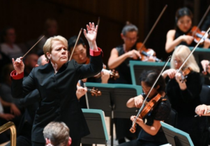 A string section of an orchestra with a white woman with short blonde hair conducting.
