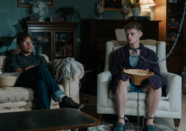 Scene from a film, the living room, darkly lit. A man watching TV with another man chained up.