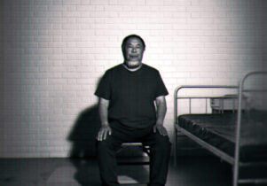 A black-and-white image of Ai Weiwei sitting upright on a small stool in a sparse, cell-like room. He faces the camera with his hands resting on his knees. Behind him is a white brick wall, and to his right there is a simple metal bed frame with a thin mattress. The lighting is stark, casting strong shadows on the wall and floor, giving the scene a stark, surveillance-like atmosphere