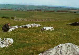 A wide countryside view shows pale limestone rocks scattered across a grassy slope, suggesting an ancient stone setting. A single person crouches beside one group of stones, while patchwork fields and low hills extend into the distance under a soft, open sky.