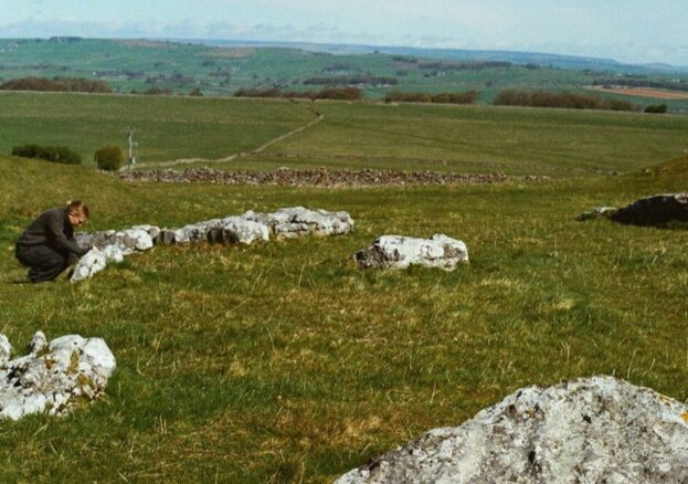 A wide countryside view shows pale limestone rocks scattered across a grassy slope, suggesting an ancient stone setting. A single person crouches beside one group of stones, while patchwork fields and low hills extend into the distance under a soft, open sky.