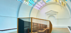 A large white room with a colourful lit ceiling above a wooden coffin shaped oversized sculpture.