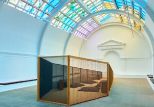 A large white room with a colourful lit ceiling above a wooden coffin shaped oversized sculpture.