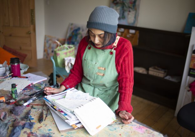 The image show the artist at work. She is wearing a red jumper/green apron looking down over the notebooks.
