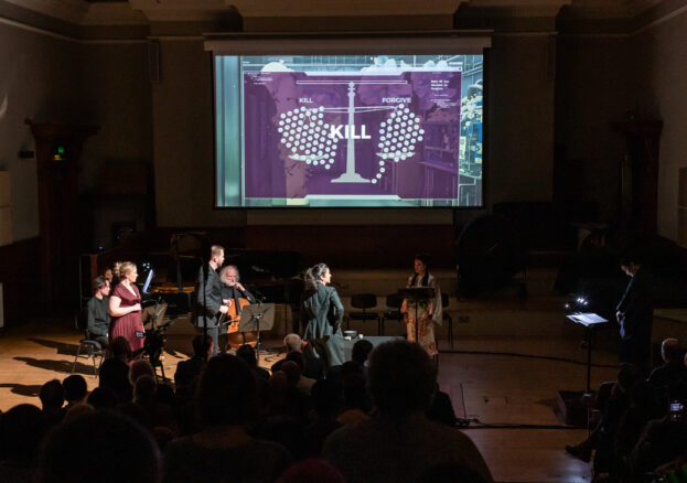 Singers and musicians on stage in a darkened concert hall, watched by a seated audience. A large screen above them shows a set of scales weighing KILL against FORGIVE, with the word KILL in large letters in the centre.